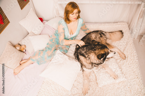 charming plus size girl with red hair in a nightgown posing with her large dog, a Malamute best friend in white bed in the bedroom