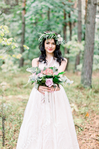 Portrait of a young beautiful bride with eucalypthus wreath on the head, with a rustic bouquet in the forest at wedding walk.