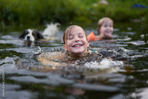 Fototapeta Two children and a dog swim in a river near the shore