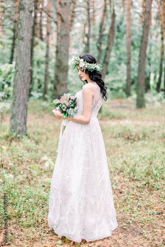 The back view of the charming brunette bride in greenery wreath on head holding the wedding bouquet during the walk in the forest.