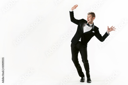 Portrait of young smiling handsome show man in tuxedo stylish black suit, studio shot dancing at white background. Businessman in jacket with bowtie