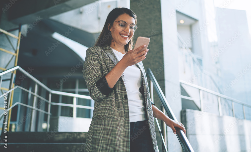 Cheerful Turkish woman dressed in elegant jacket smiling from received email with funny content, happy millennial female with modern mobile phone in hands feeling good from messaging on website