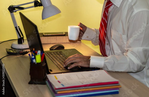 Man in suit works with laptop in office and drinks coffee. Businessman in white shirt with red tie works