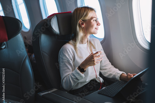 Young caucasian smiling female enjoying her comfortable flight while sitting in airplane cabin, listening to music in earphones and drinking water. Wifi internet access on board, passenger near window photo