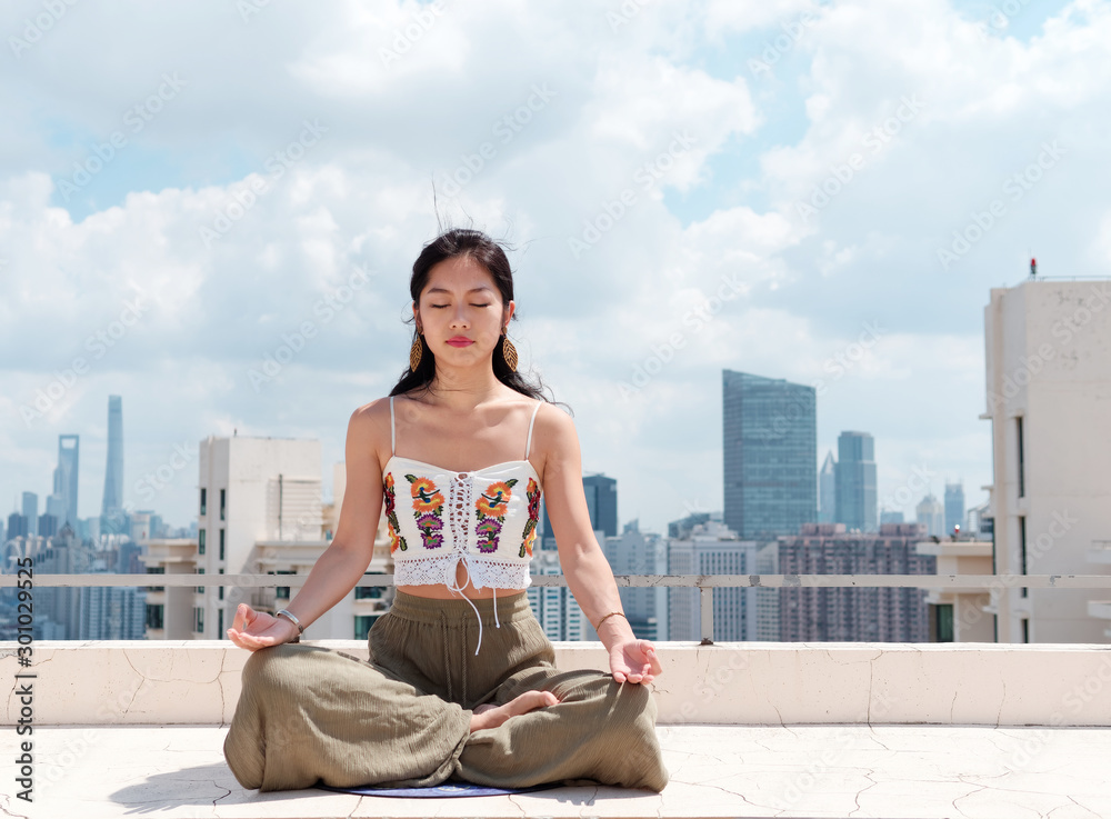 Beautiful Chinese woman wearing sport clothes practices yoga and meditates on top of building with Shanghai skyline background, young female doing yoga asana in city in sunny day.