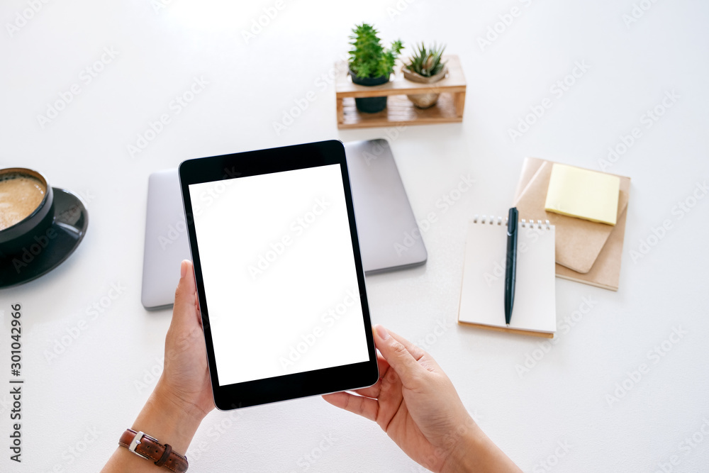 Top view mockup image of hands holding a tablet with blank white screen and laptop computer on wooden table in office Top view mockup image of hands holding a tablet with blank white screen and laptop computer on wooden table in office