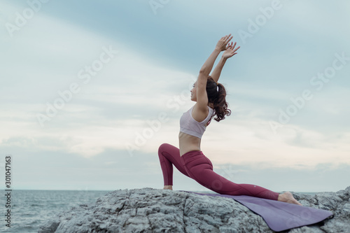 Young woman doing yoga exercise yoga master pose on the rock. Amazing yoga landscape in beautiful sky and enjoying sea view on wooden floor, concept for exercising, health care