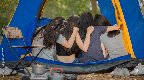 Back view, young women traveling group in tent, hugging their neck singing happily, forest in the background. Camping and travelers concept.
