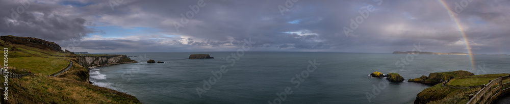 Panoramic view of a rainbow shining between rainy clouds, sea and cliffs near Ballintoy in Northern Ireland, with Sheep Island in the background.