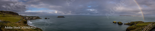 Panoramic view of a rainbow shining between rainy clouds, sea and cliffs near Ballintoy in Northern Ireland, with Sheep Island in the background.