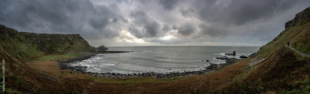Panoramic view of the pathways and the bay at the coast called Giant's Causeway, a landmark in Northern Ireland, with the cliffs that surround the place and dramatic cloudy sky.