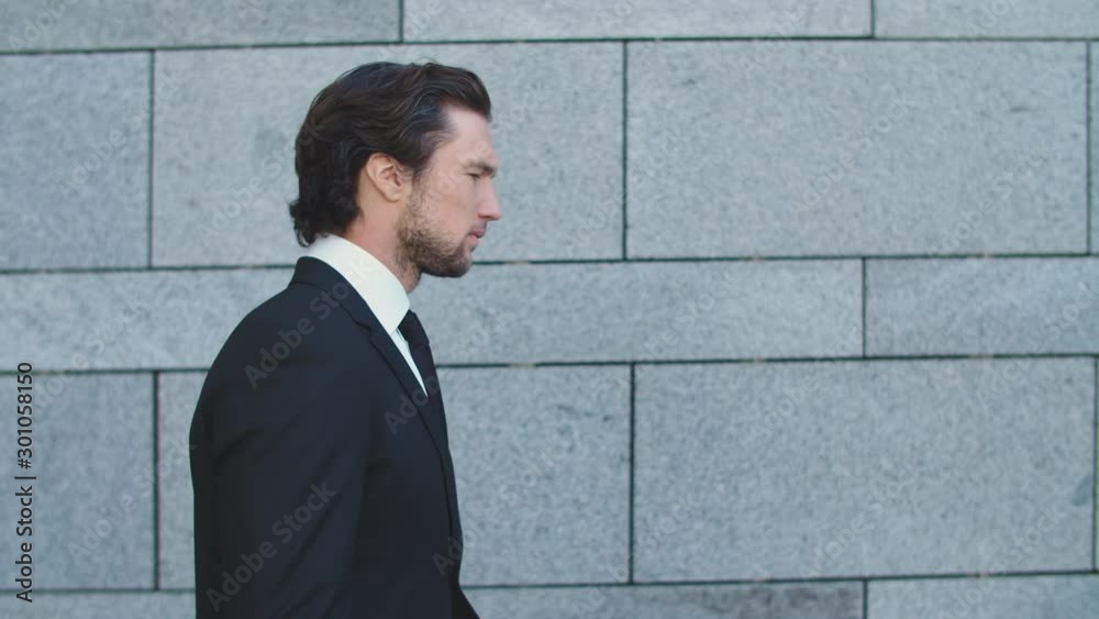 A handsome serious man in a business suit walks against a concrete wall. Guy is looking at his watch