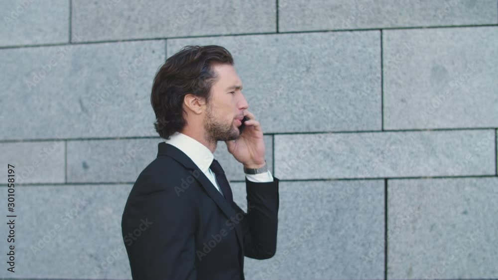 A businessman is walking down the street and talking on the phone against a gray wall. A businessman is walking down the street and talking on the phone against a gray wall.