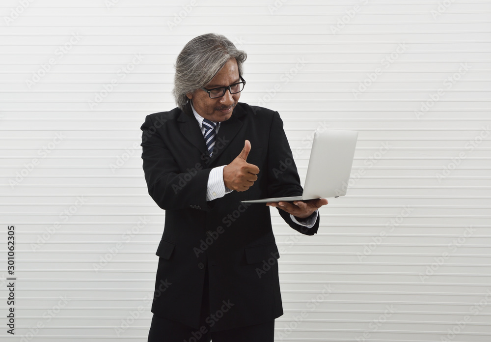 Smiling elderly asian businessman in black suit and eyeglasses holding laptop computer and making thumbs up over whit wall background, Business confident and success concept Smiling elderly asian businessman in black suit and eyeglasses holding laptop computer and making thumbs up over whit wall background, Business confident and success concept