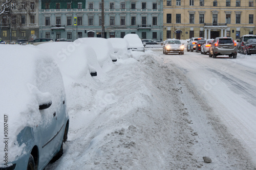 Parked cars in large snowdrifts on the side of the road in the old town and traffic on a cloudy winter morning