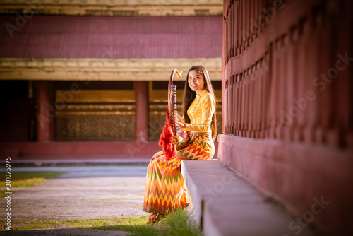 Tablou pe pânză A beautiful asian model wearing dress traditional posing smile with play musical instrument in Mandalay palace , Myanmar