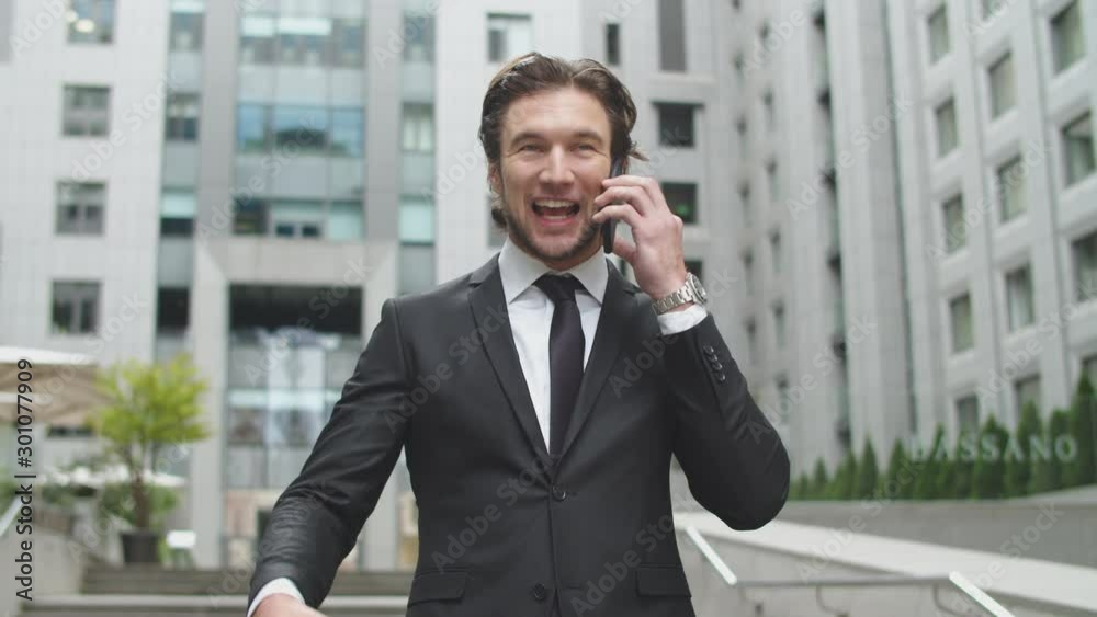 A man in a black business suit received good news during a telephone conversation. A man stands in the courtyard of a large office center. A man in a black business suit received good news during a telephone conversation. A man stands in the courtyard of a large office center.