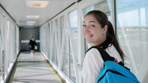 Happy young woman with backpack is going on telescopic gangway to the airplane. She turns around, looks at the camera and smiles.
