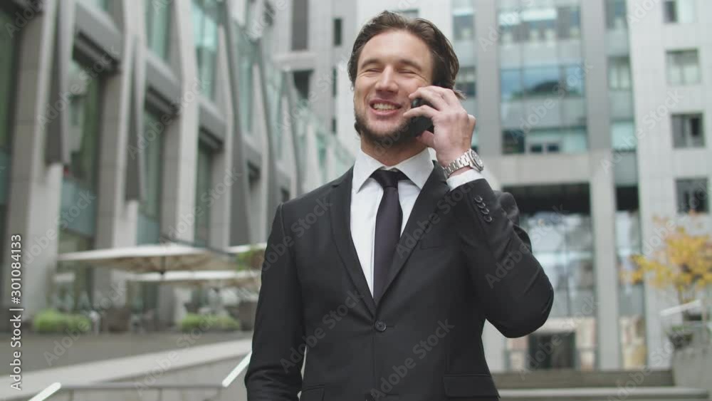 Handsome bearded man dressed in black suit talking by smartphone before modern office building Handsome bearded man dressed in black suit talking by smartphone before modern office building