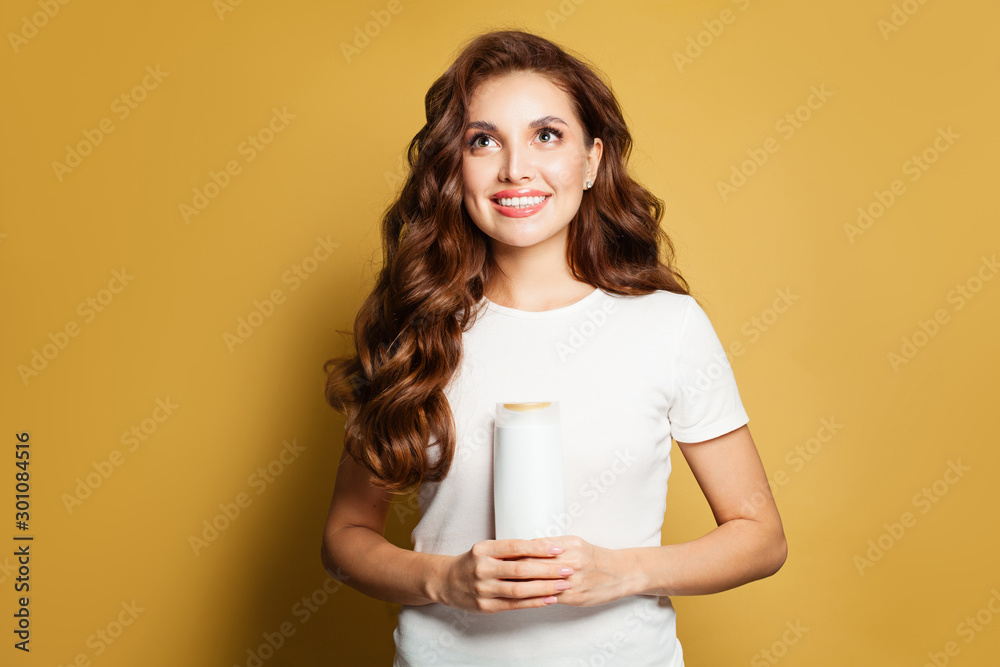 Young woman with healthy hair looking up and holding white empty shampoo tube on yellow background Young woman with healthy hair looking up and holding white empty shampoo tube on yellow background