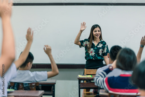 An smiling Asian female high school teacher teaches the white uniform students in the classroom by asking questions and then the students raise their hands for answers.