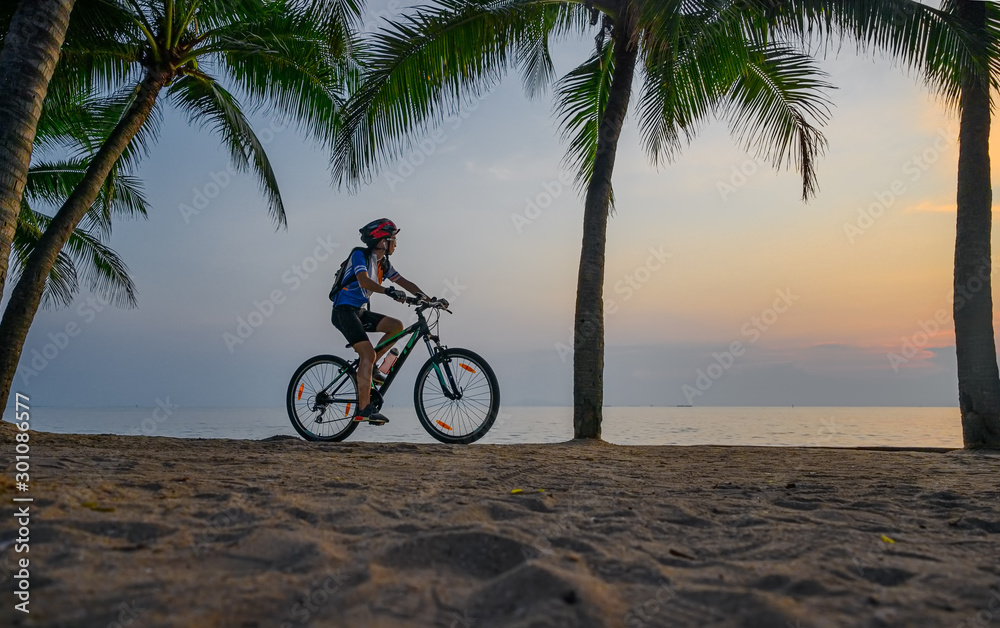 Fototapeta premium woman riding bicycle along the sea beach under coconut trees at sunset, relaxation at the end of the day.