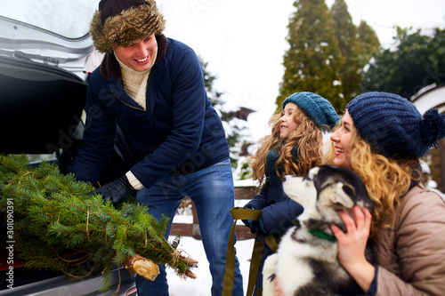 Father brought christmas tree in large trunk of SUV car. Daughter, mother and dog meet dad happily help him with holidays home decorations. Family prepares for new year together. Snowy winter outdoors photo