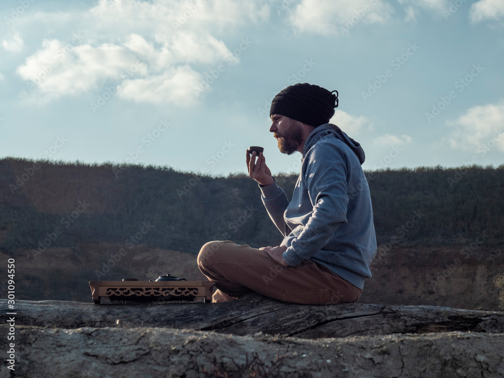 hipster man with a beard and in a knitted hat drinks tea from a bowl on a background of mountains and sky. chinese tea ceremony