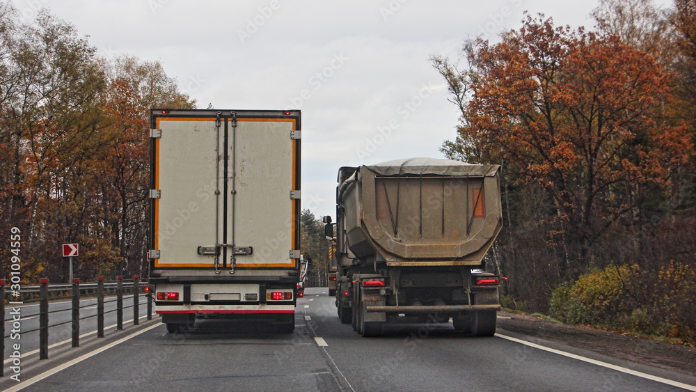 Semi truck with van trailer overtake flatbed dump truck on autumn country highway road, rear view