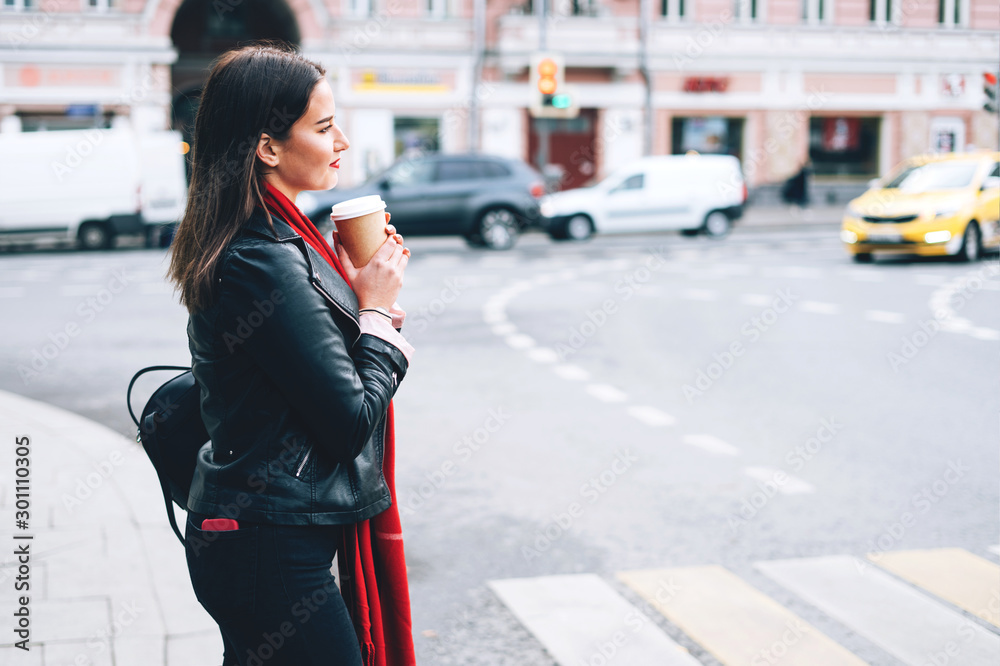 A beautiful brunette woman standing at the street in a black leather jacket and a red scarf and hold in hand a hot drink.