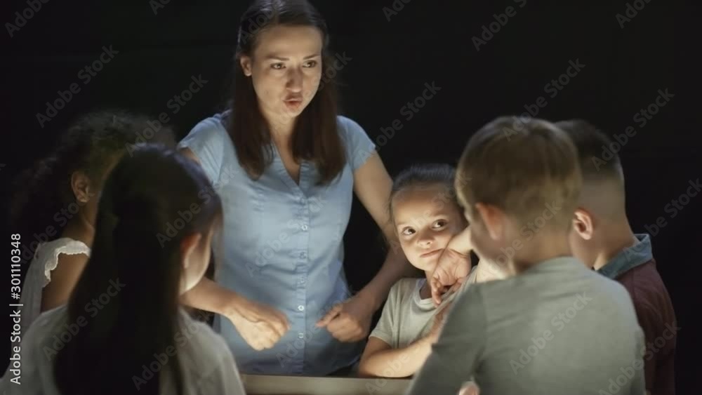 Tracking shot of cheerful young woman teaching group of kids how to make sand art animation in dark classroom, then looking at camera and smiling Tracking shot of cheerful young woman teaching group of kids how to make sand art animation in dark classroom, then looking at camera and smiling