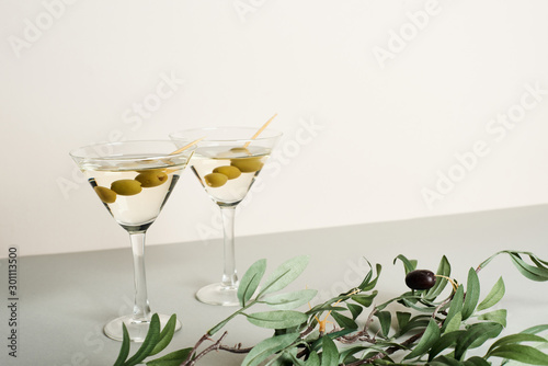 Cocktails in martini glasses with olive branch on grey surface, isolated on white