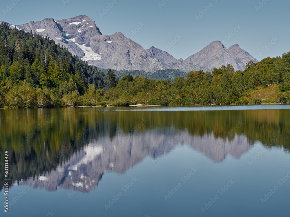 Panoramic view of Kardyvach lake in sunny morning among Caucasus mountains; mirror water surface reflecting autumn highland landscape, rocky peaks and overgrown steep slopes of Agepsta mountain ridges