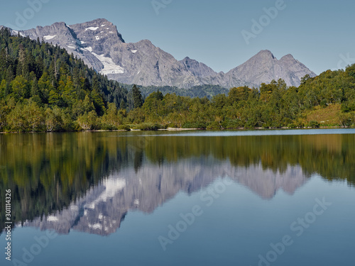 Panoramic view of Kardyvach lake in sunny morning among Caucasus mountains  mirror water surface reflecting autumn highland landscape  rocky peaks and overgrown steep slopes of Agepsta mountain ridges