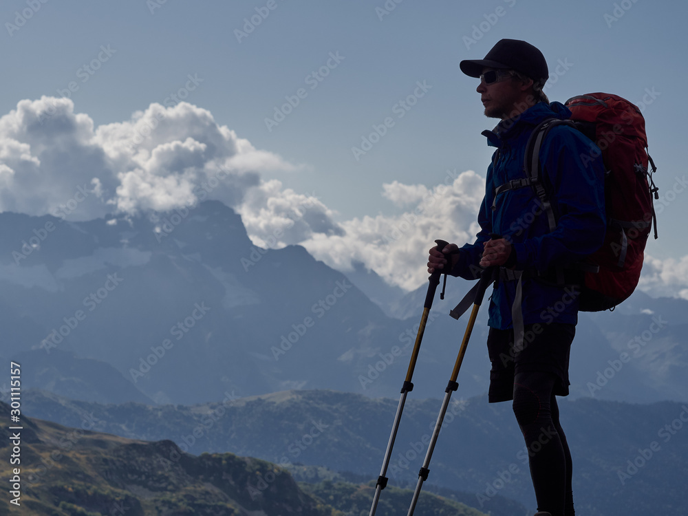 Silhouette of young hiker standing on cliff edge, view in full height; brave man in with climbing equipment enjoying a freedom of ascent; mountain ranges and cloudy sky on background