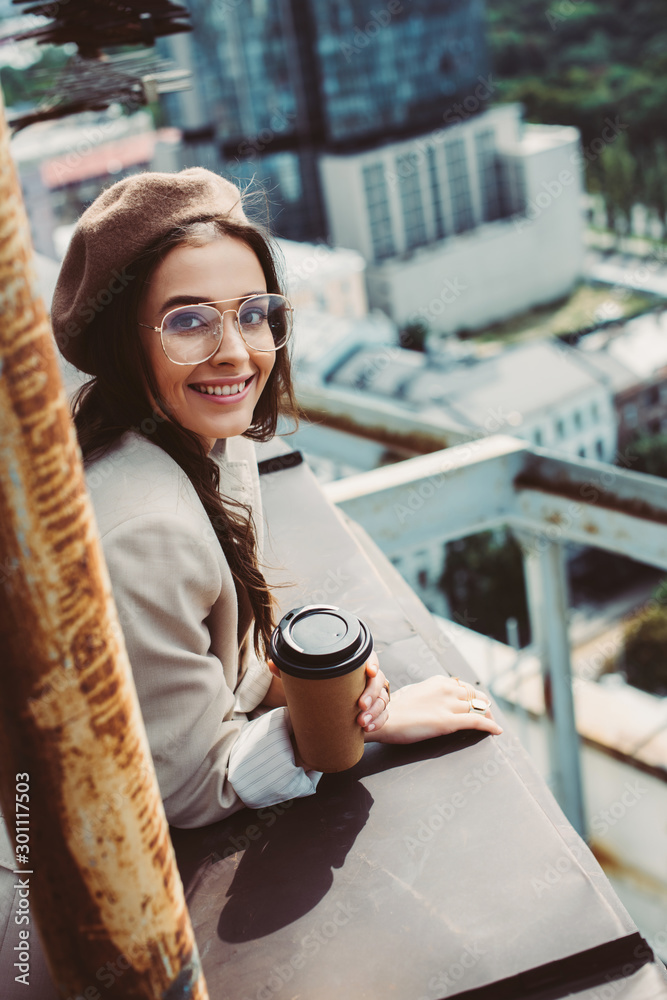 smiling fashionable girl in beige suit and beret posing on roof with coffee to go