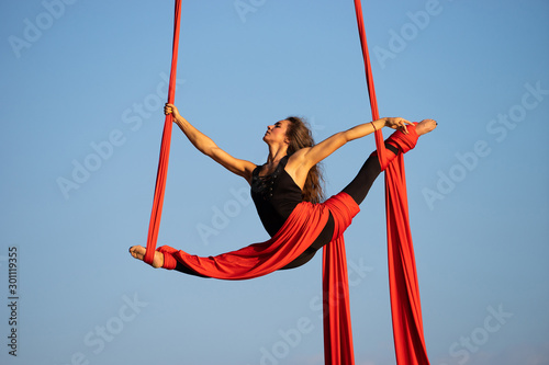 Beautiful and flexible female circus artist dancing with aerial silk on a sky background