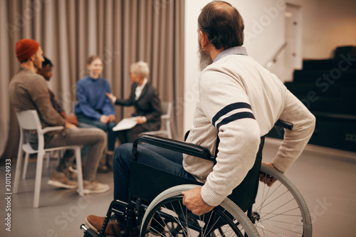 Rear view of senior man sitting in wheelchair and looking at people sitting and talking during therapy class