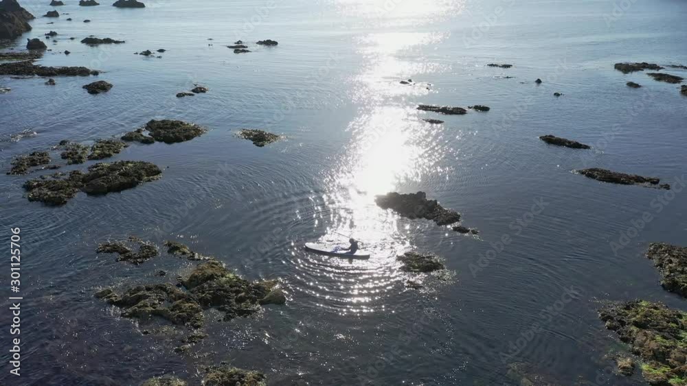 Aerial view of climbing onto Stand Up Paddle board on calm water with sun reflecting along the rocky coastline in the South of Ireland on a calm day Aerial view of climbing onto Stand Up Paddle board on calm water with sun reflecting along the rocky coastline in the South of Ireland on a calm day