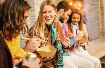 Happy friends eating street food outdoor - Young trendy people having fun together drinking and laughing around downtown streets - City lifestyle and party concept - Focus on blond girl face
