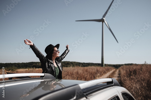 Happy Traveling Girl Enjoying a Car Trip on the Field Road with Electric Wind Turbine Power Generator on the Background
