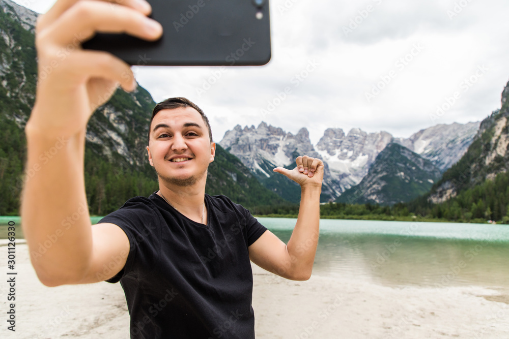 Young man in high mountain near alpine lake taking selfie portrait with beautiful landscape. Traveling man using mobile phone to take selfie in summer