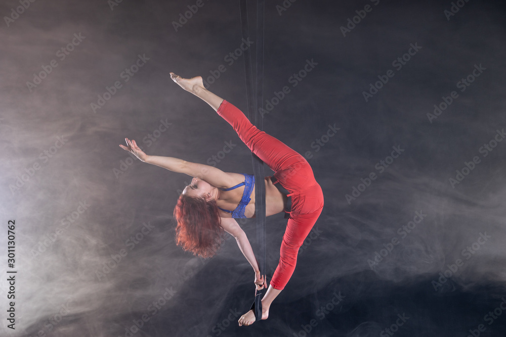 Female athletic, sexy and flexible aerial circus artist with redhead on aerial straps on black background Female athletic, sexy and flexible aerial circus artist with redhead on aerial straps on black background
