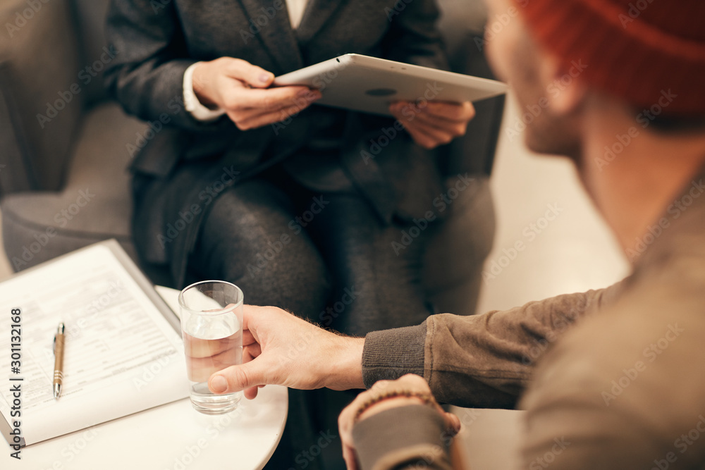 Obraz premium Rear view of young man taking glass of water from the table while talking to businesswoman at meeting