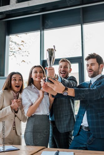 happy multicultural businesswomen and businessmen looking at trophy in office