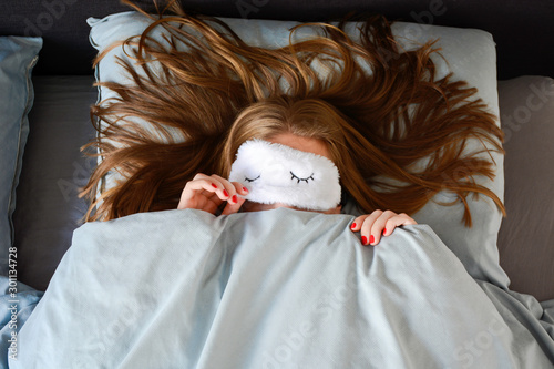 Young woman with long hair and a red manicure is sleeping in bed with sleep mask and peeking from underneath the sleeping mask