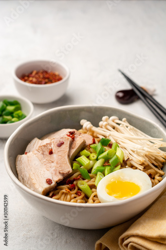 Asian soup ramen with noodles, pork, spring onion, enoki mushrooms and boiled egg in bowl on concrete background. Selective focus.