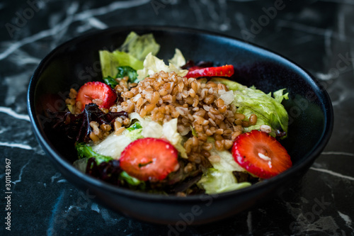 Buckwheat Salad with Strawberry Slices and Green Leaves in Black Bowl on Dark Granite Surface.
