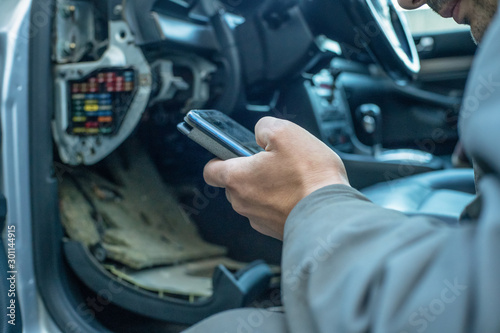 male mechanic looks at the phone information about the cause of the breakdown of the car sitting next to the car. Concept of car repair