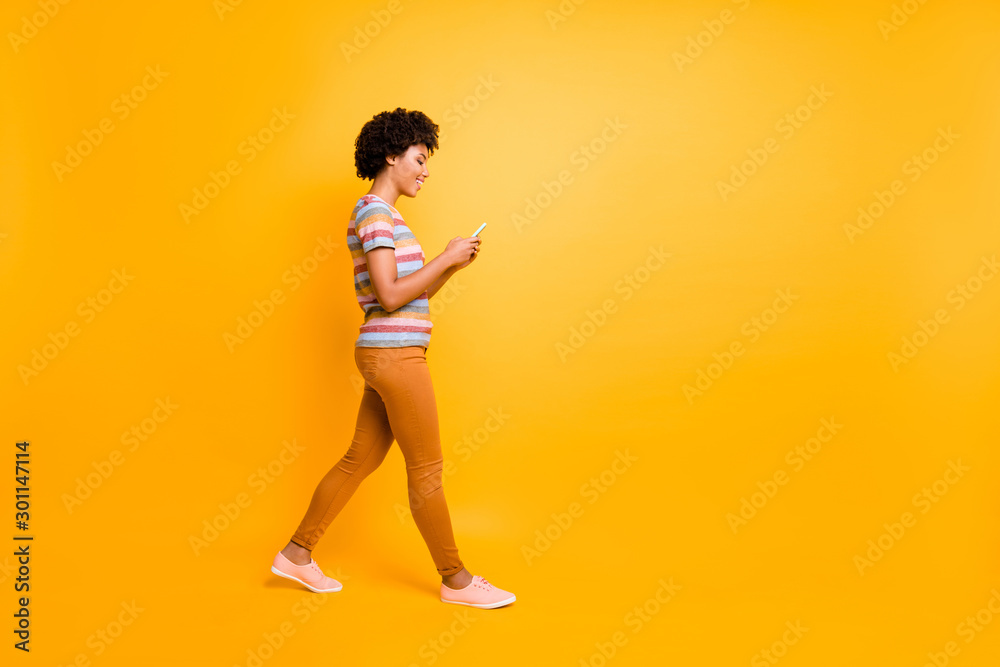 Full length profile side photo of positive dark skin brown hair girl use smartphone read social media feednews go walk travel summer wear striped t-shirt outfit isolated bright color background Full length profile side photo of positive dark skin brown hair girl use smartphone read social media feednews go walk travel summer wear striped t-shirt outfit isolated bright color background